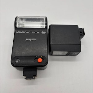 Peut inclure: Un flash électronique pour appareil photo Agfatronic 261 CB noir avec une lentille transparente, un bouton d'alimentation orange vif et une étiquette computer. À côté se trouve un composant flash rectangulaire noir avec une lentille.