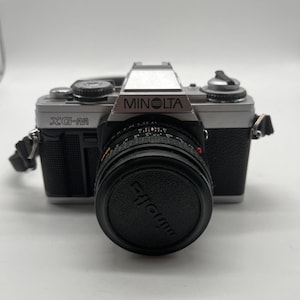 Könnte beinhalten: Eine silber-schwarze Minolta XG-M 35mm Filmkamera mit einem schwarzen Objektivdeckel, der das Minolta-Logo trägt. Das Kameragehäuse hat einen strukturierten schwarzen Griff, eine silberne Oberseite mit Einstellrädern und ein Objektiv.