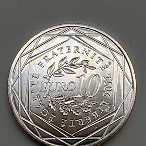 Peut inclure: Une pièce de 10 euros argentée de 2011 présentant un motif central composé d'une branche d'olivier et de feuilles de chêne. L'inscription LIBERTE EGALITE FRATERNITE entoure le motif, avec le chiffre 10 et le mot EURO au centre.