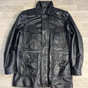 Könnte beinhalten: Eine schwarze Lederjacke mit klassischem Kragen, Knopfleiste vorne und langen Ärmeln. Das Kleidungsstück verfügt über zwei schräge Brusttaschen mit Metallreißverschlüssen sowie zwei große aufgesetzte Taschen mit geknöpften Klappen und Nähten.