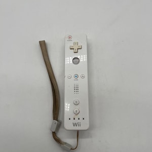 Può includere: Un controller per videogiochi bianco con pulsante di accensione, tasto direzionale, pulsanti A, 1 e 2, tasto home e griglia dell'altoparlante. La parola Wii è stampata in basso. È attaccato un cinturino da polso beige con cursore in plastica.