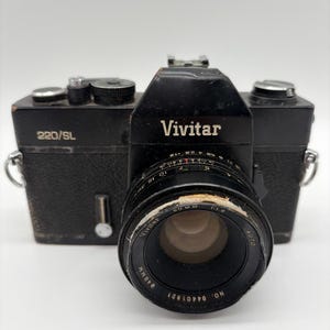 May include: A black Vivitar 220/SL 35mm film camera with a textured body. The lens is marked Vivitar 50mm 1:1.8 Auto, with a 49mm filter thread. The camera features a silver shutter button, a film advance lever, and a hot shoe mount.