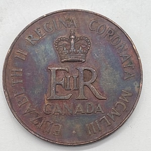 Op de afbeelding: Een ronde Canadese herdenkingsmunt met een centrale kroon boven het koninklijk monogram ER II. De tekst ELIZABETH II REGINA CORONATA MCMLIII en CANADA staat rondom de rand gegraveerd. De munt heeft een donkere, verkleurde metalen afwerking.