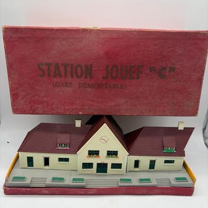 May include: A model train station building with cream-colored walls, dark green doors and windows, and a dark red roof. The structure is displayed in front of its original red cardboard box labeled STATION JOUEF C (GARE DEMONTABLE).