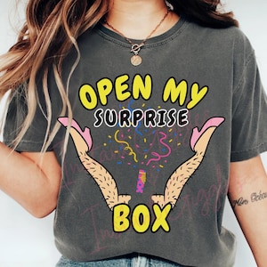 Könnte beinhalten: Dunkelgraues T-Shirt mit dem Text "OPEN MY SURPRISE BOX" in Gelb und Schwarz. Das Design zeigt Cartoon-Beine in rosa High Heels mit Konfetti und Luftschlangen.