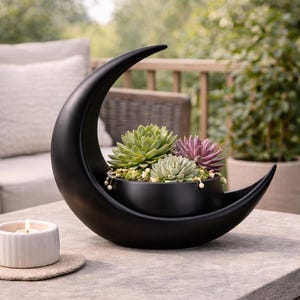 Könnte beinhalten: Ein schwarzer, halbmondförmiger Pflanztopf mit mehreren Sukkulenten. Der Pflanztopf steht auf einer Steinoberfläche, mit einer kleinen Kerze und einem Untersetzer im Vordergrund. Der Hintergrund zeigt eine Terrasse.
