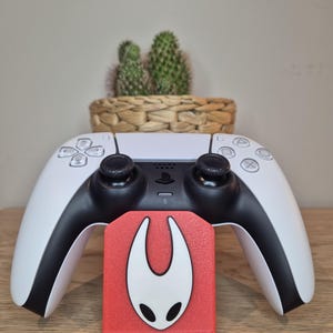 Pode incluir: Um controle PlayStation 5 branco e preto com um gráfico vermelho e branco. O controle está em frente a um pequeno cacto em uma cesta de vime. O gráfico é um rosto estilizado.