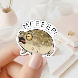 May include: A sticker featuring a cartoon frog with yellow eyes and an open mouth, expressing surprise. The frog is tan and beige, with the word "MEEEEP!" in white text above it. The sticker is held by a person.