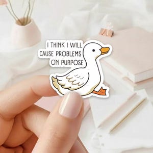 May include: A white duck sticker with an orange beak and feet, and the text "I THINK I WILL CAUSE PROBLEMS ON PURPOSE". The sticker is held between fingers against a soft background.