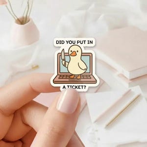 May include: A sticker featuring a cartoon duck holding a knife, standing in front of a laptop. The text on the sticker reads "DID YOU PUT IN A TICKET?" The sticker is held by a person.