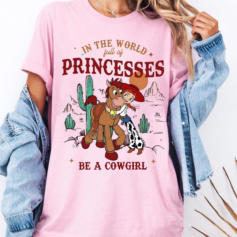 May include: Pink t-shirt with a graphic featuring a cartoon cowgirl and horse in a desert scene. Text reads "In the world full of princesses, be a cowgirl." The design includes cacti and a setting sun. The shirt has short sleeves.