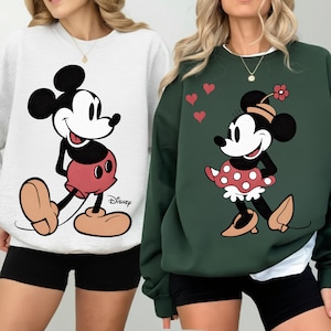 Könnte beinhalten: Zwei Sweatshirts mit Disney-Figuren. Eines ist weiß mit Mickey Mouse in roten Shorts. Das andere ist dunkelgrün mit Minnie Mouse in einem gepunkteten Rock. Beide Sweatshirts haben schwarze Shorts.