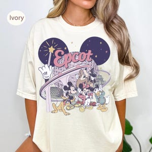 May include: Ivory-colored t-shirt featuring a graphic of Disney characters in front of the Epcot ball. The design includes Mickey Mouse, Minnie Mouse, Donald Duck, Goofy, and Pluto. The word "Epcot" is written in a pink script font.