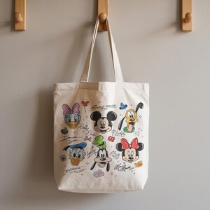 May include: Off-white canvas tote bag featuring colorful illustrations of Disney characters, including Mickey Mouse, Minnie Mouse, Donald Duck, and Pluto. The bag has two handles and is hanging on a wooden hook.