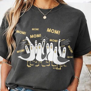 May include: Dark gray t-shirt featuring a cartoon illustration of seagulls with the word "MOM!" in gold lettering above each bird. The seagulls are white and gray with yellow feet.