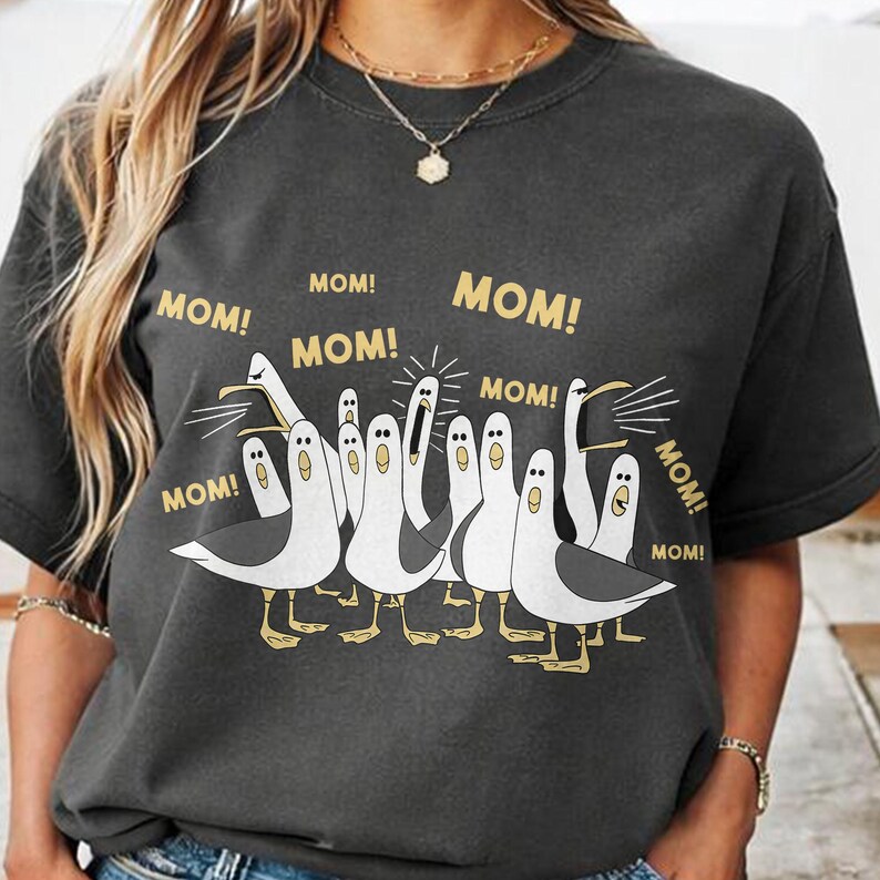 May include: Dark gray t-shirt featuring a cartoon illustration of seagulls with the word "MOM!" in gold lettering above each bird. The seagulls are white and gray with yellow feet.