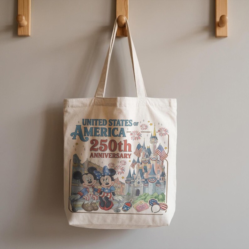 May include: Cream-colored tote bag featuring Mickey and Minnie Mouse in patriotic attire, with a castle and fireworks illustration. The bag has the text "UNITED STATES OF AMERICA 250th ANNIVERSARY" in blue and red.