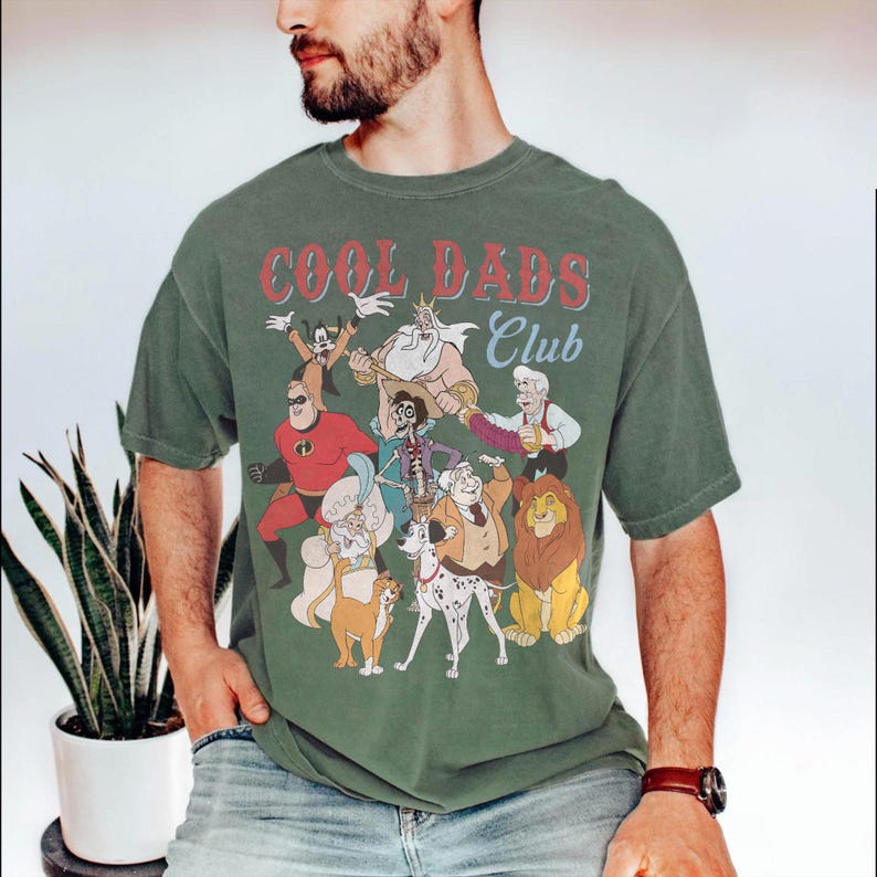 K&ouml;nnte beinhalten: Olivgr&uuml;nes T-Shirt mit einer Grafik verschiedener Zeichentrickfiguren und dem Text "COOL DADS CLUB" in Rot und Wei&szlig;. Das Shirt hat einen Vintage-Look.