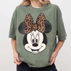 K&ouml;nnte beinhalten: Olivgr&uuml;nes T-Shirt mit gro&szlig;em Minnie Mouse-Motiv. Das Design zeigt einen schwarzen Kopf und Ohren, ein wei&szlig;es Gesicht und eine Schleife mit Leopardenmuster. Das T-Shirt ist im l&auml;ssigen Stil gehalten.