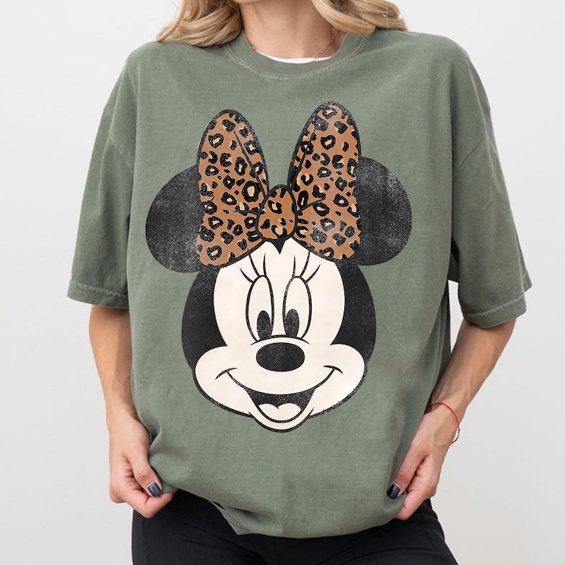 K&ouml;nnte beinhalten: Olivgr&uuml;nes T-Shirt mit gro&szlig;em Minnie Mouse-Motiv. Das Design zeigt einen schwarzen Kopf und Ohren, ein wei&szlig;es Gesicht und eine Schleife mit Leopardenmuster. Das T-Shirt ist im l&auml;ssigen Stil gehalten.