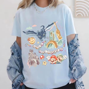 Op de afbeelding: Lichtblauw T-shirt met een kleurrijk grafisch ontwerp met een draak, een kasteel en de tekst "EPIC UNIVERSE". Het ontwerp bevat ook Mario en Luigi, een toverstaf en een lachende zon.