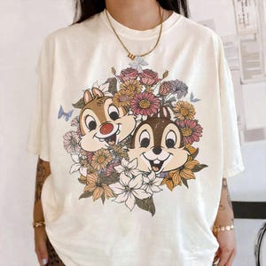 May include: Cream-colored t-shirt featuring a graphic of Chip and Dale surrounded by a floral arrangement in shades of pink, orange, and white. The design includes butterflies and is printed on a short-sleeved tee.