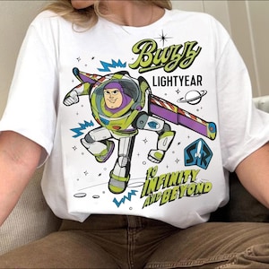 May include: White t-shirt featuring a colorful graphic of Buzz Lightyear from Toy Story. The design includes the text "Buzz Lightyear" and "To Infinity and Beyond" with a space-themed illustration. The shirt is a casual wear item.