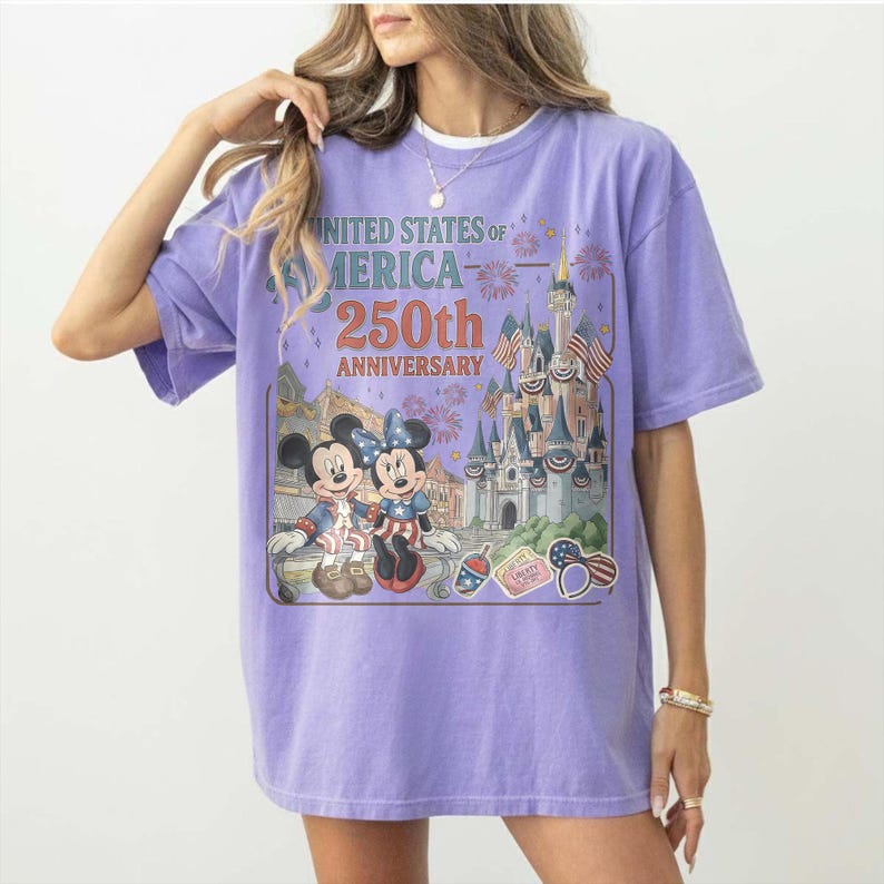 May include: A lilac-colored t-shirt featuring a graphic commemorating the 250th anniversary of the United States of America. The design includes Mickey and Minnie Mouse, a castle, fireworks, and patriotic imagery.