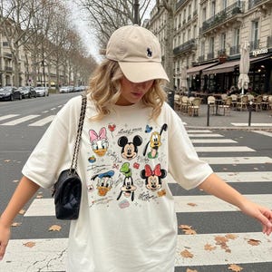 May include: Cream-colored t-shirt featuring a colorful graphic of Disney characters, including Mickey Mouse, Minnie Mouse, and Donald Duck. The shirt has short sleeves and a crew neck. A beige baseball cap with a small logo is worn. A black shoulder bag with a chain strap is also visible.