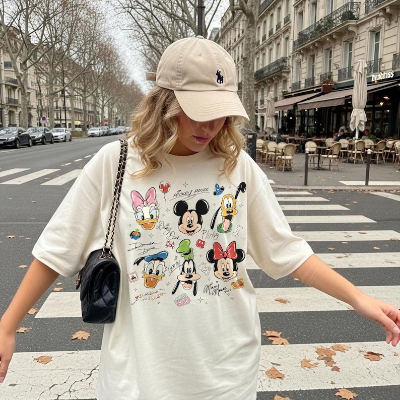 May include: Cream-colored t-shirt featuring a colorful graphic of Disney characters, including Mickey Mouse, Minnie Mouse, and Donald Duck. The shirt has short sleeves and a crew neck. A beige baseball cap with a small logo is worn. A black shoulder bag with a chain strap is also visible.