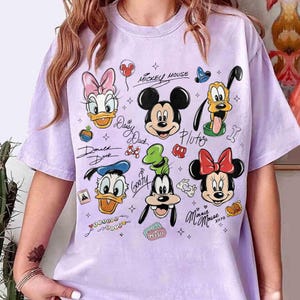 May include: A light purple t-shirt featuring various Disney cartoon characters, including Mickey Mouse, Minnie Mouse, Donald Duck, Daisy Duck, Goofy, and Pluto. The shirt has a crew neck and short sleeves. The characters are in a cartoon style.