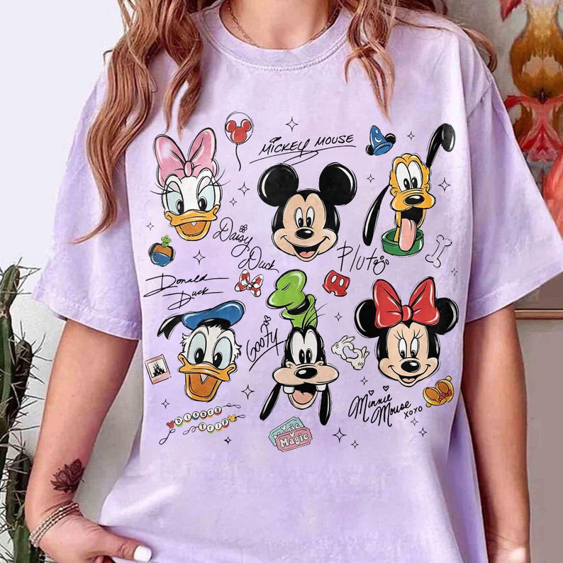 May include: A light purple t-shirt featuring various Disney cartoon characters, including Mickey Mouse, Minnie Mouse, Donald Duck, Daisy Duck, Goofy, and Pluto. The shirt has a crew neck and short sleeves. The characters are in a cartoon style.