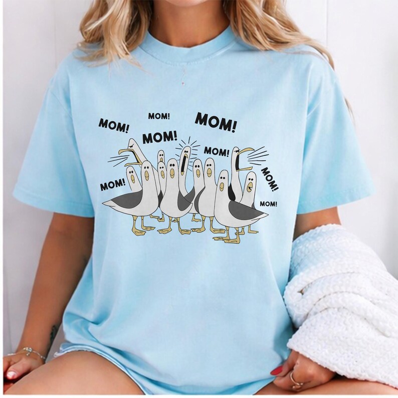 May include: Light blue t-shirt featuring a cartoon illustration of seagulls with the word "MOM!" in speech bubbles. The seagulls are in a row, with black and white bodies and yellow feet. The text "MOM!" is repeated around the seagulls.