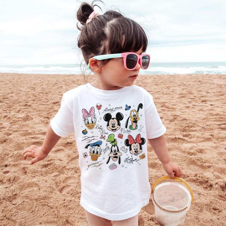 May include: A white t-shirt featuring colorful cartoon character graphics, including Mickey Mouse, Minnie Mouse, and Donald Duck. The shirt is worn by a young child on a sandy beach, with the ocean visible in the background.