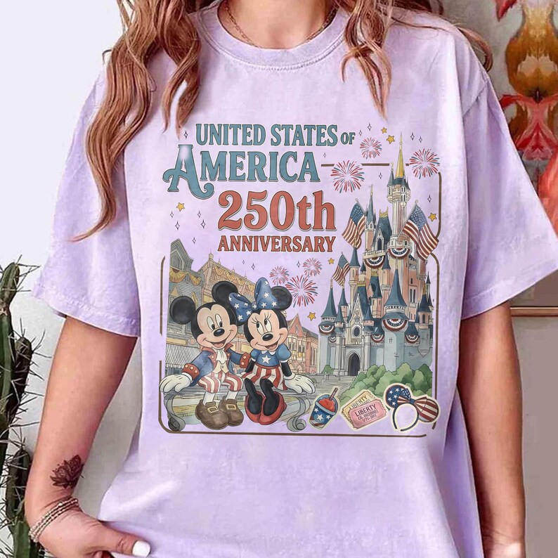 May include: A light purple t-shirt featuring a graphic celebrating the United States of America's 250th Anniversary. The graphic includes Mickey and Minnie Mouse, a castle, fireworks, and the text "UNITED STATES OF AMERICA - 250th ANNIVERSARY."