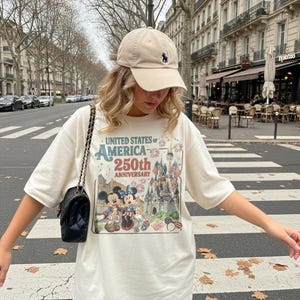 May include: Cream-colored t-shirt with a graphic celebrating the 250th anniversary of the United States of America. The design includes Mickey and Minnie Mouse, a castle, and American flags. A beige baseball cap with a dark blue logo is worn.