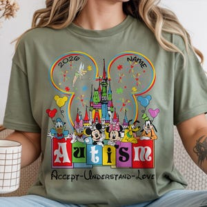 May include: Olive green t-shirt featuring a colorful graphic with Disney characters, a castle, and the word "Autism." The design includes the text "Accept-Understand-Love" and the year "2026."