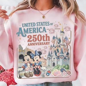 May include: Pink sweatshirt featuring a graphic celebrating the 250th anniversary of the United States of America. The design includes Mickey and Minnie Mouse in patriotic attire, a castle, fireworks, and the text "UNITED STATES OF AMERICA 250th ANNIVERSARY."