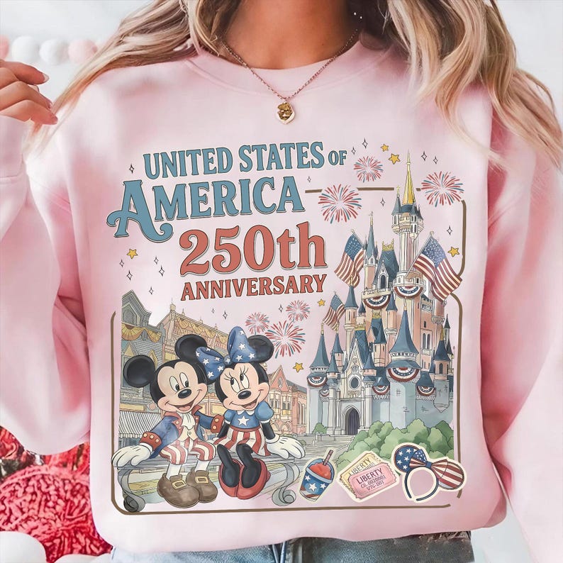 May include: Pink sweatshirt featuring a graphic celebrating the 250th anniversary of the United States of America. The design includes Mickey and Minnie Mouse in patriotic attire, a castle, fireworks, and the text "UNITED STATES OF AMERICA 250th ANNIVERSARY."