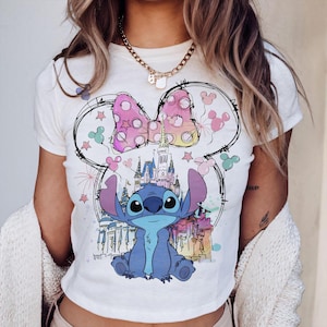 May include: White t-shirt featuring a cartoon illustration of Stitch from Lilo & Stitch. The design includes a pink polka-dot bow, a castle, and Mickey Mouse silhouettes. The shirt has a crew neck and short sleeves.