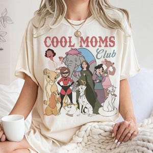 May include: Cream-colored t-shirt featuring the text "COOL MOMS Club" above a graphic of cartoon characters. The graphic includes an elephant, a lion cub, a dalmatian, and other animated figures. The shirt has short sleeves.