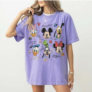 May include: A lilac-colored t-shirt featuring a graphic of Mickey Mouse and friends. The design includes images of Mickey, Minnie, Donald, Daisy, Goofy, and Pluto, along with their signatures. The shirt has short sleeves and a crew neck.