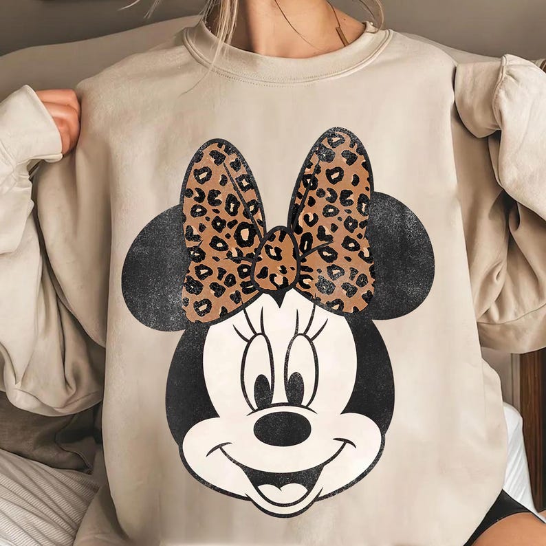 K&ouml;nnte beinhalten: Beiges Sweatshirt mit Minnie Mouse-Grafik. Die Grafik zeigt ein schwarz-wei&szlig;es Minnie Mouse-Gesicht mit einer Schleife im braunen Leopardenmuster. Das Sweatshirt hat lange &Auml;rmel und einen Rundhalsausschnitt.