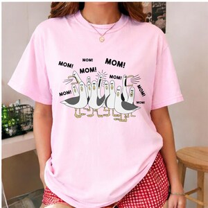 May include: Pink t-shirt featuring a cartoon illustration of seagulls with the word "MOM!" repeated around them. The seagulls are white with gray wings and yellow feet. The shirt is a light pink color.