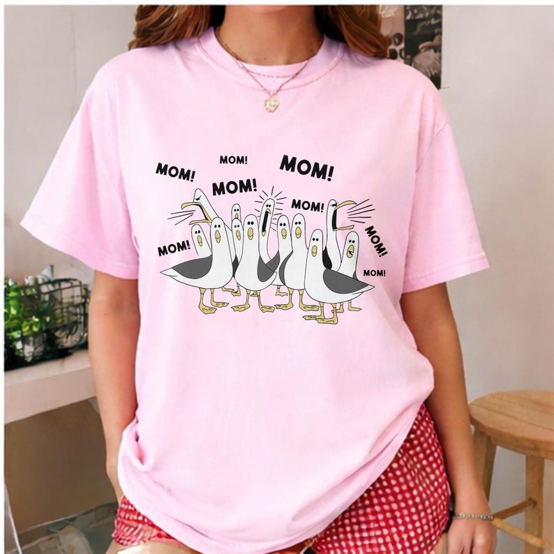 May include: Pink t-shirt featuring a cartoon illustration of seagulls with the word "MOM!" repeated around them. The seagulls are white with gray wings and yellow feet. The shirt is a light pink color.