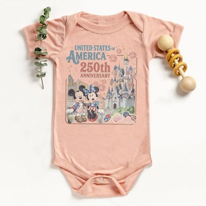 May include: A light pink baby onesie featuring a graphic celebrating the United States of America's 250th Anniversary. The graphic includes Mickey and Minnie Mouse, a castle, and patriotic imagery. A wooden rattle is on the right.