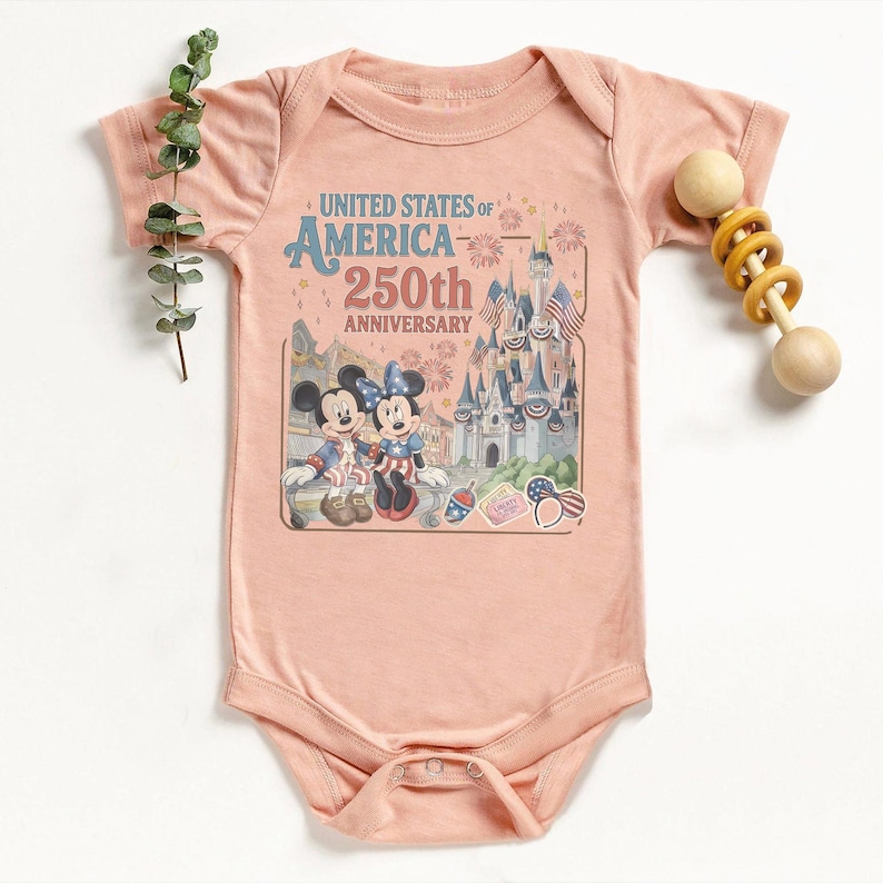 May include: A light pink baby onesie featuring a graphic celebrating the United States of America's 250th Anniversary. The graphic includes Mickey and Minnie Mouse, a castle, and patriotic imagery. A wooden rattle is on the right.