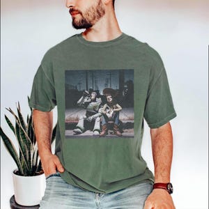 May include: Green t-shirt featuring a graphic of two animated characters sitting on a curb. One character is a space ranger, and the other is a cowboy. The graphic is in a vintage style.