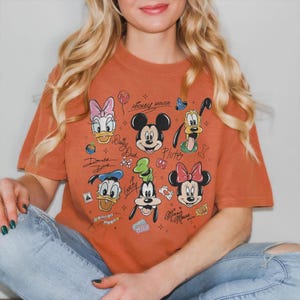 May include: Burnt orange t-shirt featuring a graphic of Mickey Mouse, Minnie Mouse, Donald Duck, Daisy Duck, Goofy, and Pluto. The design includes character signatures and small icons. The shirt is a relaxed fit.
