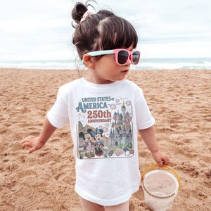 May include: A white t-shirt featuring a graphic commemorating the 250th anniversary of the United States of America. The design includes the text "United States of America 250th Anniversary" along with images of Mickey and Minnie Mouse, a castle, and fireworks.